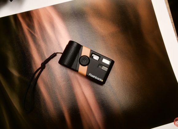 A small black and orange film camera with a wrist strap on a warm, abstract brown background.