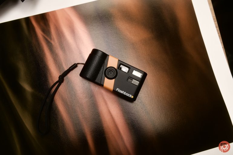 A small black and orange film camera with a wrist strap on a warm, abstract brown background.