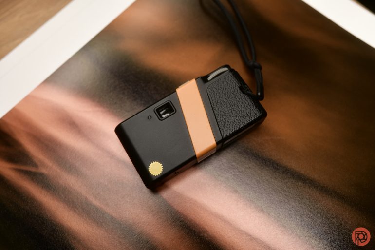 A compact black camera with a tan strap lies on a blurred brown and peach background.