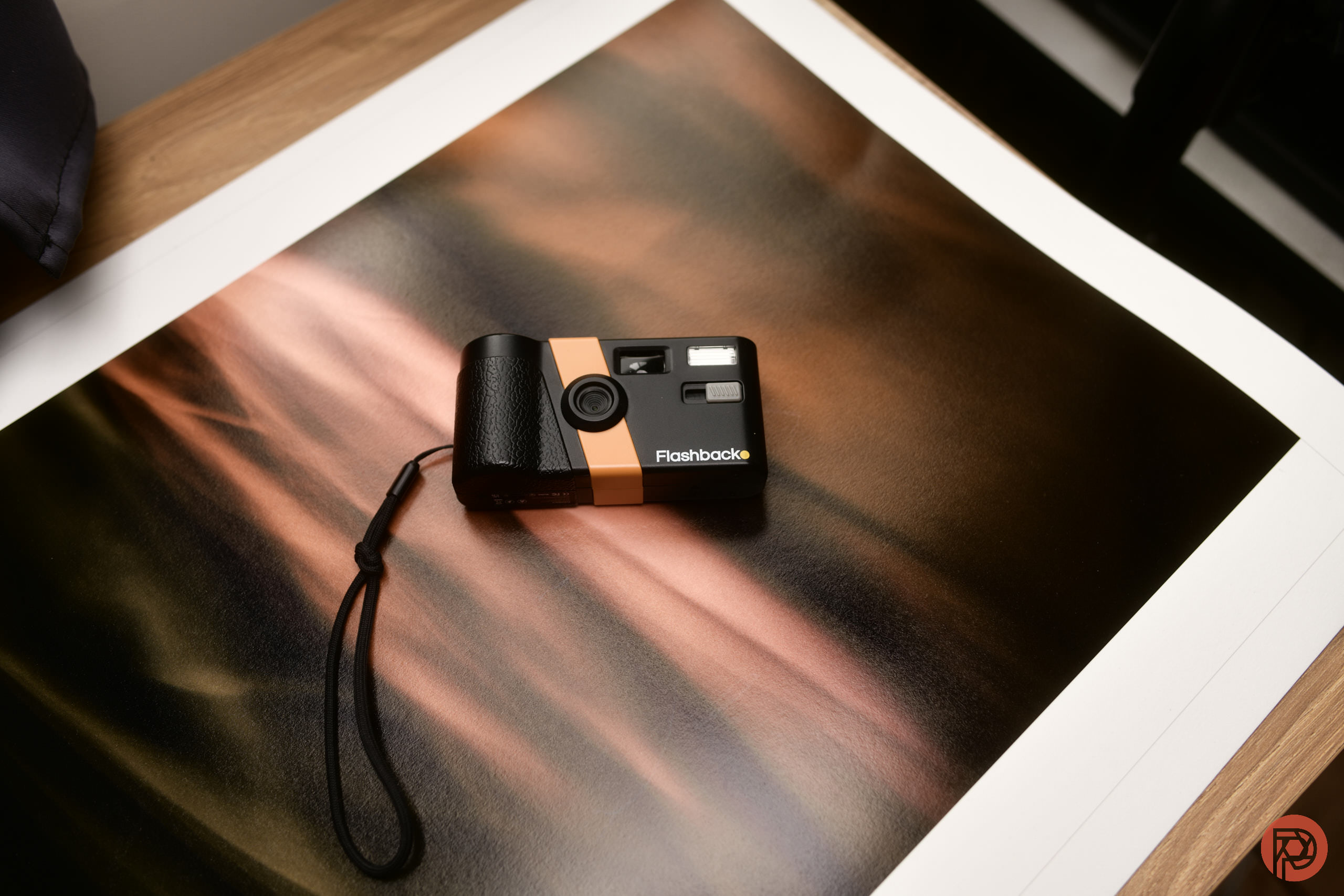 The 3 Best Cheap Compact Cameras for Travel in 2026 A black and orange film camera with a wrist strap rests on an abstract print with warm tones.