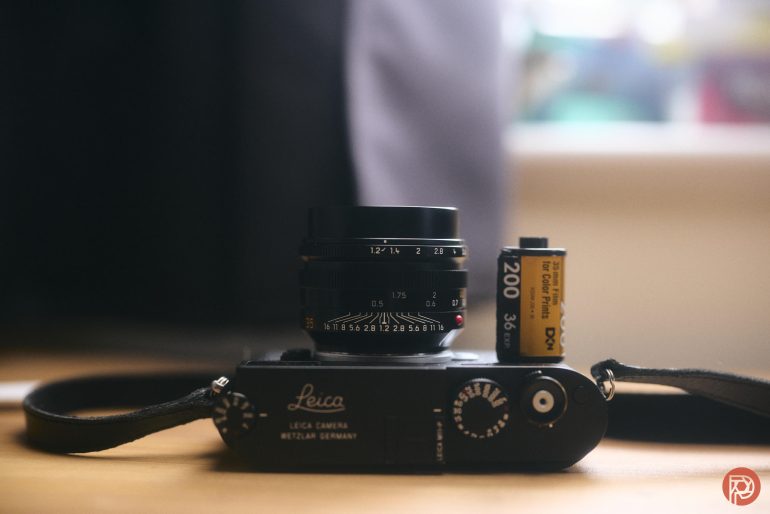 A Leica camera and a 200 ISO roll of 35mm film placed on a wooden surface indoors.