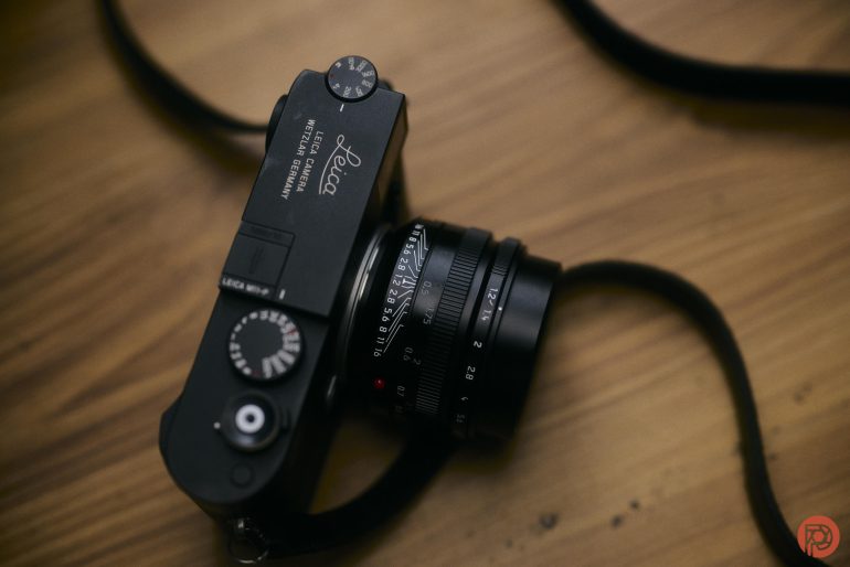Black Leica camera with a lens, placed on a wooden surface with a strap partially visible.