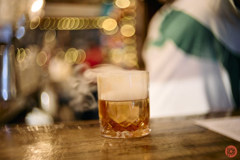 A glass of amber whiskey with smoke on top sits on a wooden bar, with blurred lights in the background.