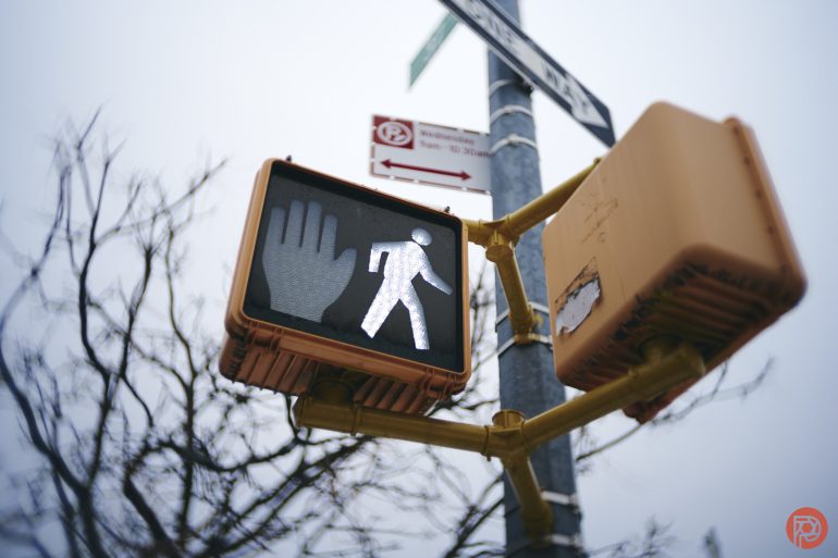 A crosswalk signal shows both the white walking figure and the raised hand to stop, with a street sign above.