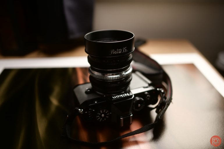A Nikon camera with a Petzval 85mm lens rests on a blurred surface in soft, warm lighting.