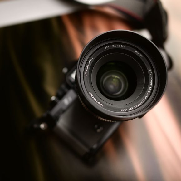 A close-up of a camera lens facing upward, with a blurred background and camera strap visible.
