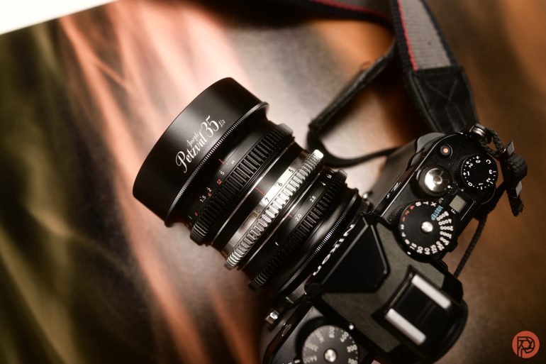 Close-up of a black digital camera with a large lens labeled Perar 35 on a blurred brown background.