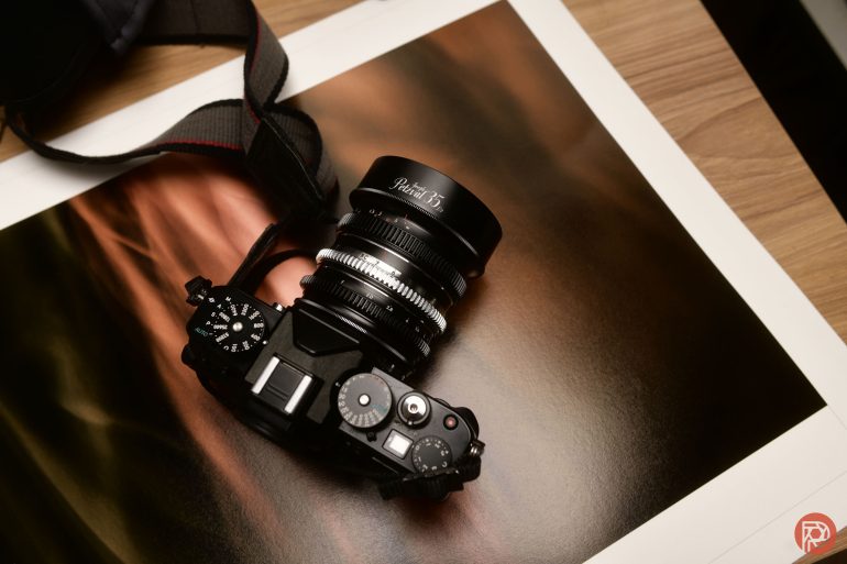 A black camera with a Petzval lens rests on a printed photo on a wooden surface.