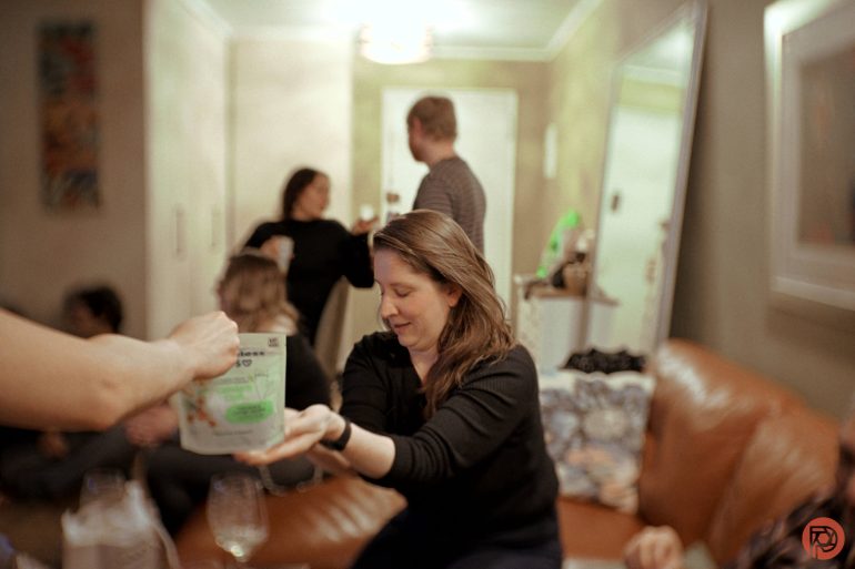 A woman on a couch reaches for a drink pouch while others gather and talk in a cozy living room.