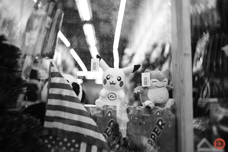 Stuffed Pikachu and another plush toy sit in a store window behind an American flag, in black and white.
