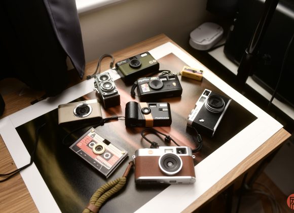 Several vintage and modern compact cameras are arranged on a wooden desk under soft natural light.