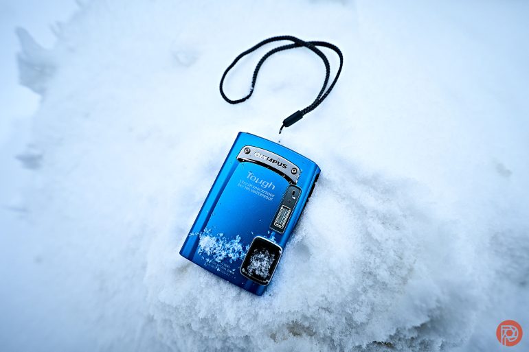 A blue Olympus Tough camera with a strap lies on snow, partially covered with snow in the corner.