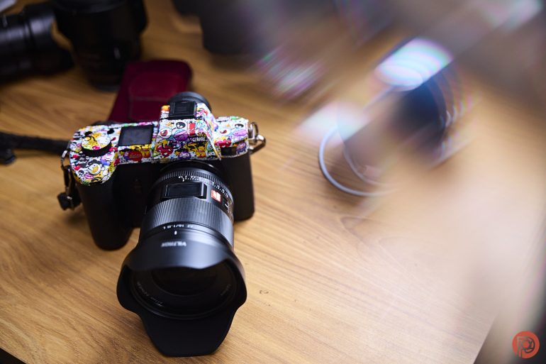 A camera with a colorful strap sits on a wooden table, surrounded by blurred objects.