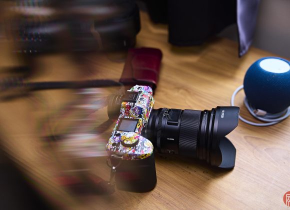 A colorful camera with stickers rests on a wooden desk near a blue smart speaker and wallet.