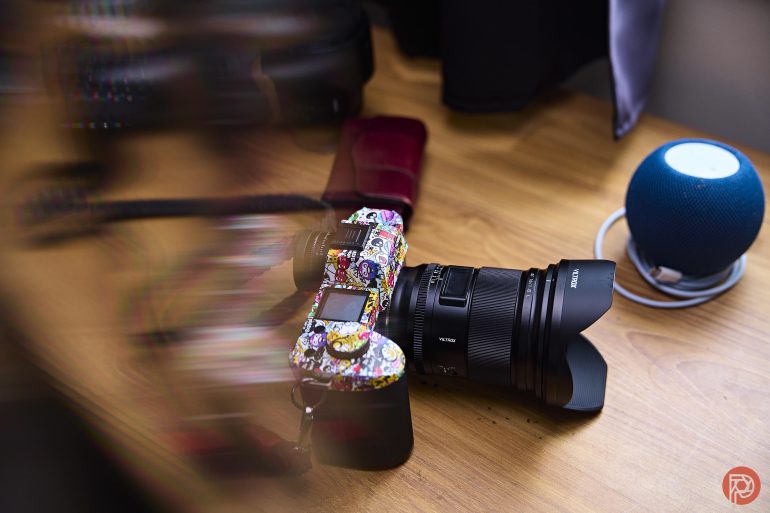 A colorful camera with stickers rests on a wooden desk near a blue smart speaker and wallet.