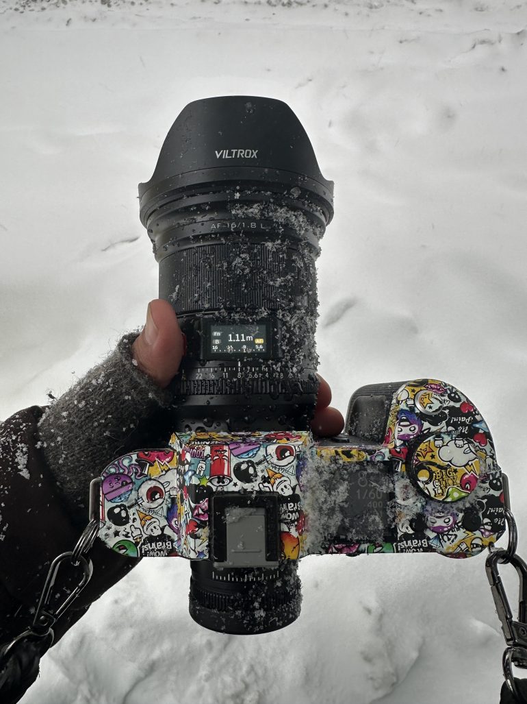 A gloved hand holds a snow-dusted, sticker-covered camera in a snowy outdoor setting.