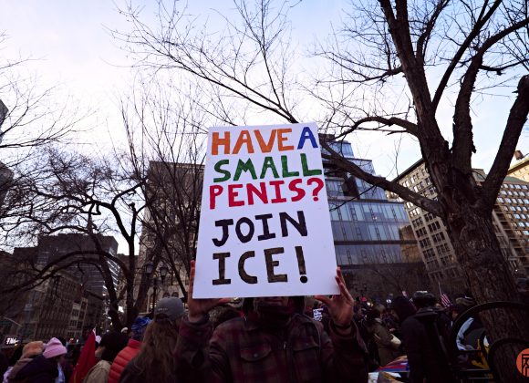 Person holds a protest sign reading Have a small penis? Join ICE! in a city park with trees and buildings.