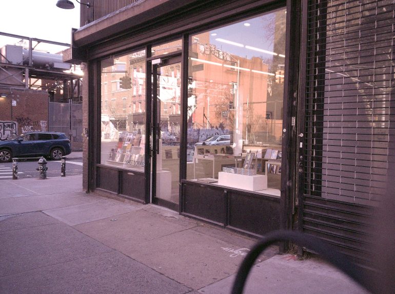 A bookstore with large glass windows on a city street, with books displayed inside and cars parked outside.