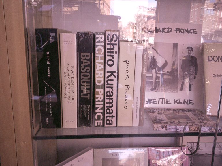 Books about art and artists are displayed upright and stacked on a glass shelf in a bookstore.