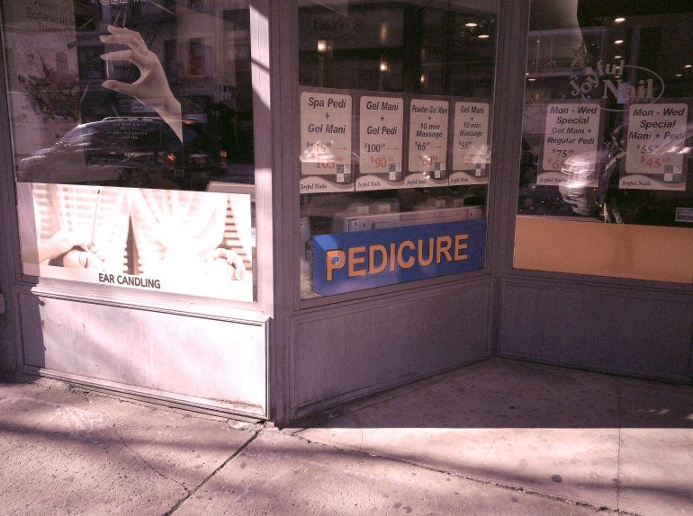 Nail salon window with PEDICURE sign and various manicure and spa service prices displayed.