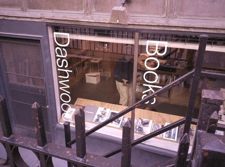 View through a window of Dashwood Books, showing shelves, tables, and a person inside the bookstore.
