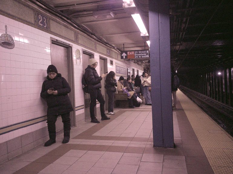People wait on a subway platform, some standing and using phones, under fluorescent lights.