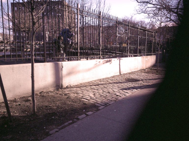 A cobblestone path runs beside a metal fence with bare trees and a person with a backpack visible in the background.