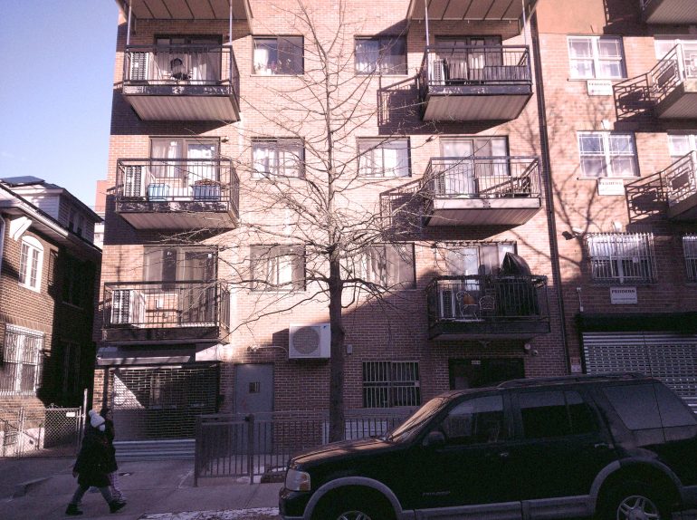 A four-story brick apartment building with balconies, a tree in front, and a parked SUV.