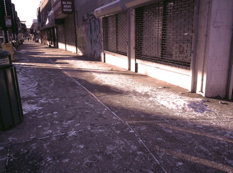 A sunlit city sidewalk with patches of ice and closed storefronts, early in the morning.