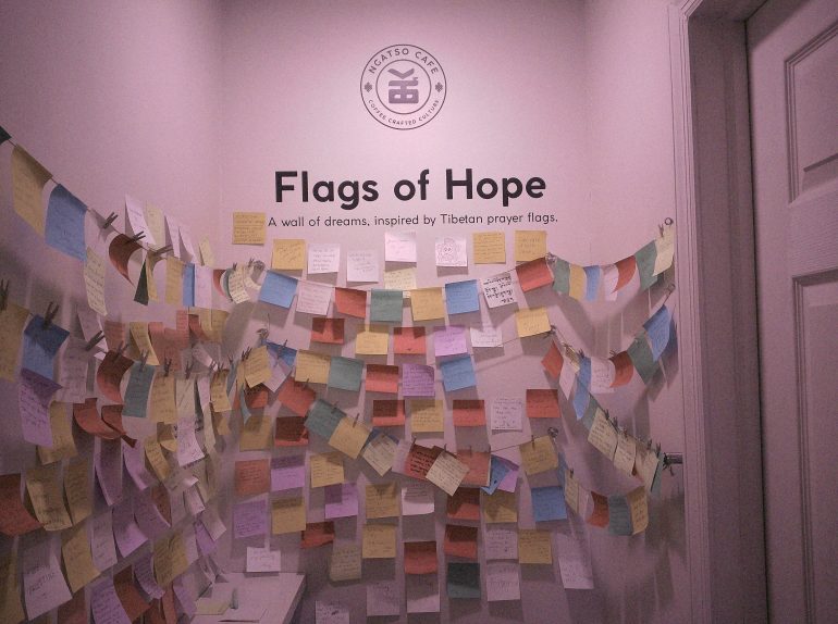 Wall filled with colorful notes labeled Flags of Hope at a cafe, inspired by Tibetan prayer flags.