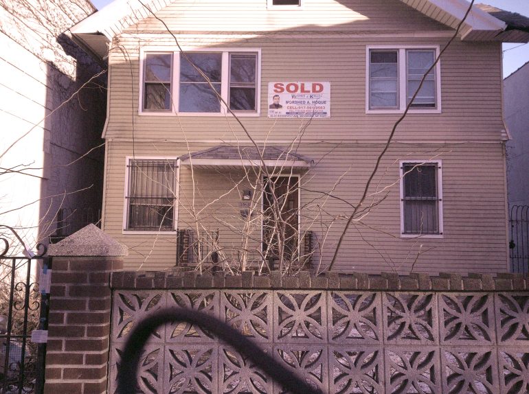 A beige two-story house with a SOLD sign above the front porch, behind a brick and patterned fence.