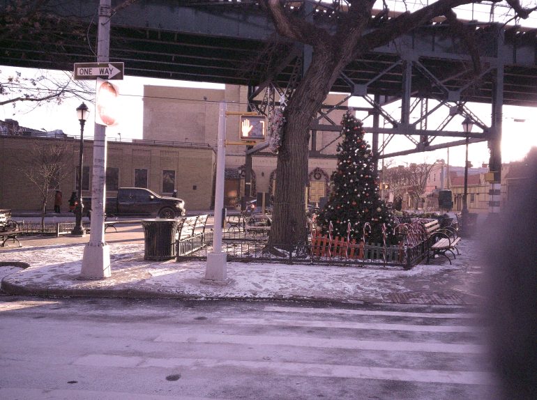 Snowy street corner with a decorated Christmas tree, benches, and a one-way sign under an overpass.