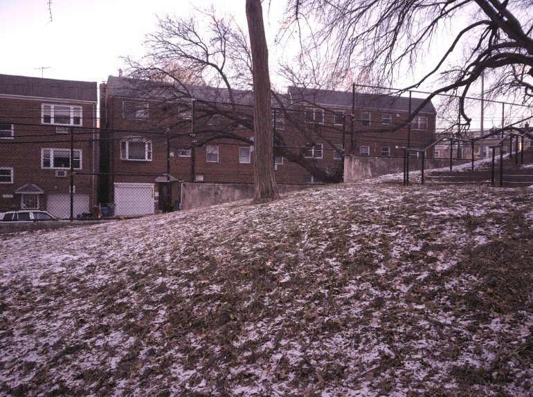 A lightly snow-dusted hill with bare trees, in front of brick apartment buildings and a black chain-link fence.