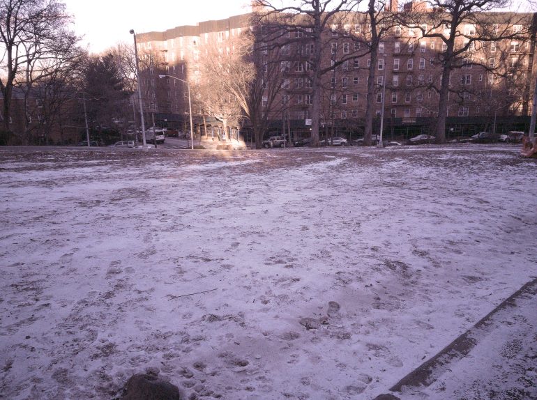 A park covered in light snow with leafless trees and apartment buildings in the background.