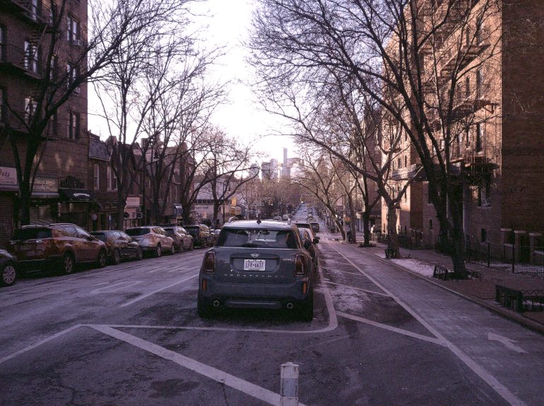 A city street lined with parked cars and leafless trees, with tall buildings in the distant background.