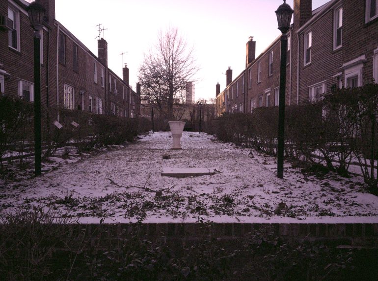 A snowy courtyard between brick buildings with a bare tree and a white pedestal in the center at dusk.