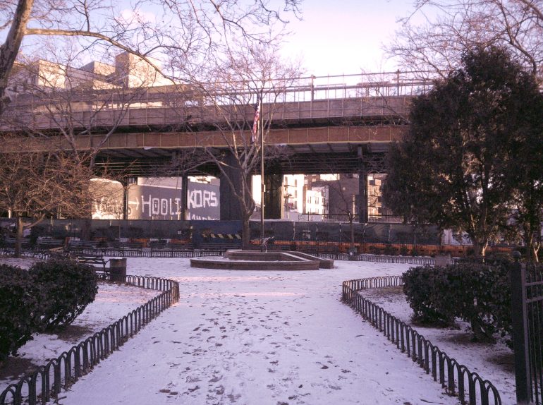 A snowy park path leads to a round fountain with an overpass and graffiti in the background.