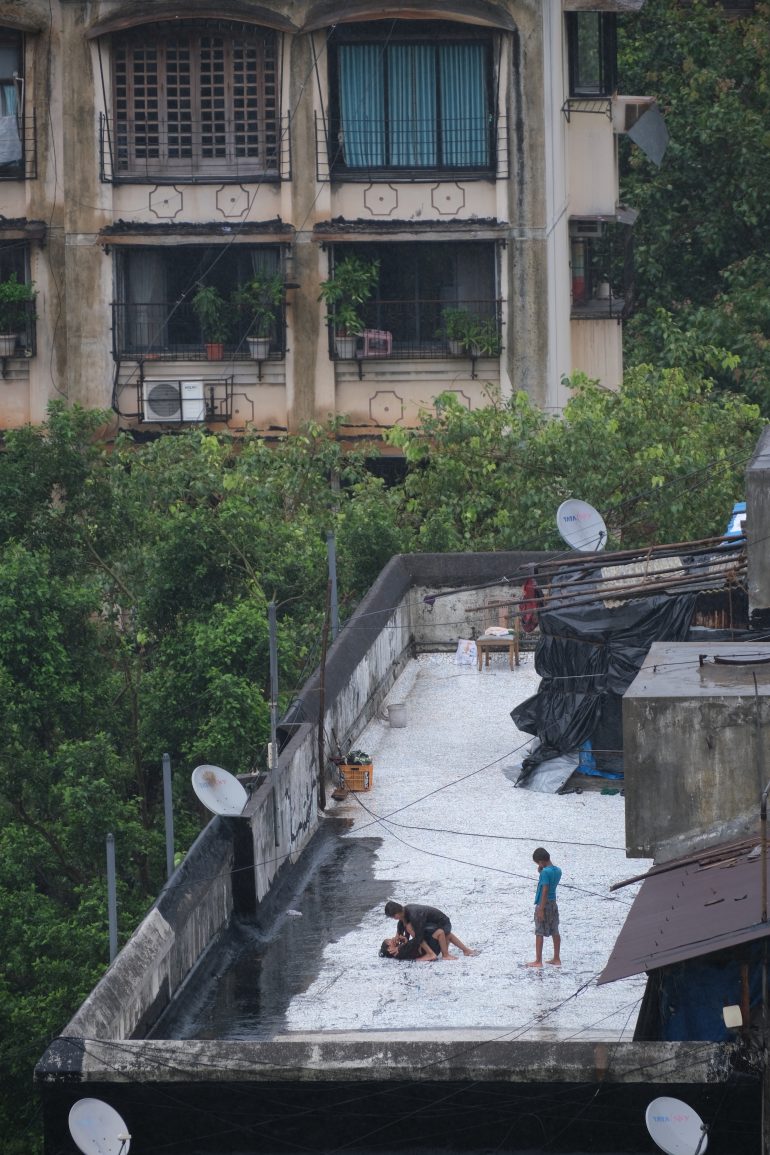Children play on a rooftop covered with water and foam, with apartment buildings and greenery in the background.