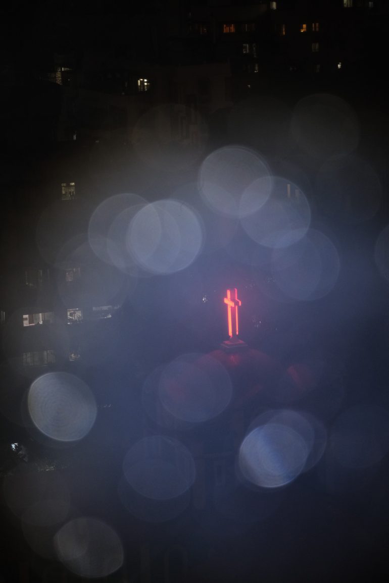 A glowing red neon cross shines through fog and bokeh lights against a dark urban background at night.