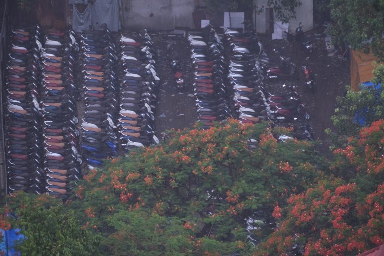 Rows of parked scooters behind trees with red-orange flowers, seen from above on a rainy day.