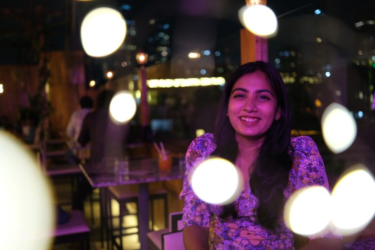 A smiling woman in a floral dress sits indoors at night, with bright blurred lights in the foreground.