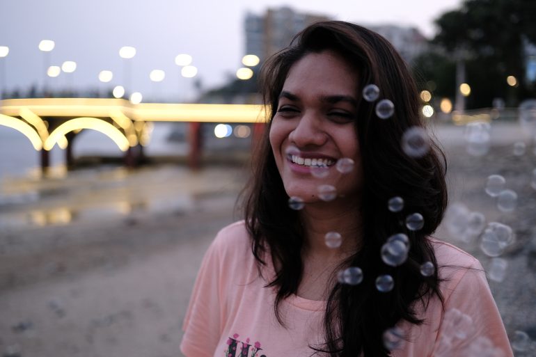 Smiling woman standing on a beach at dusk with bubbles floating in the foreground.