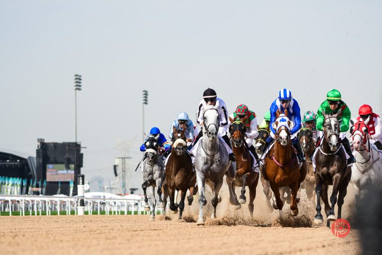 A group of jockeys riding horses race down a dirt track, kicking up dust under a clear sky.