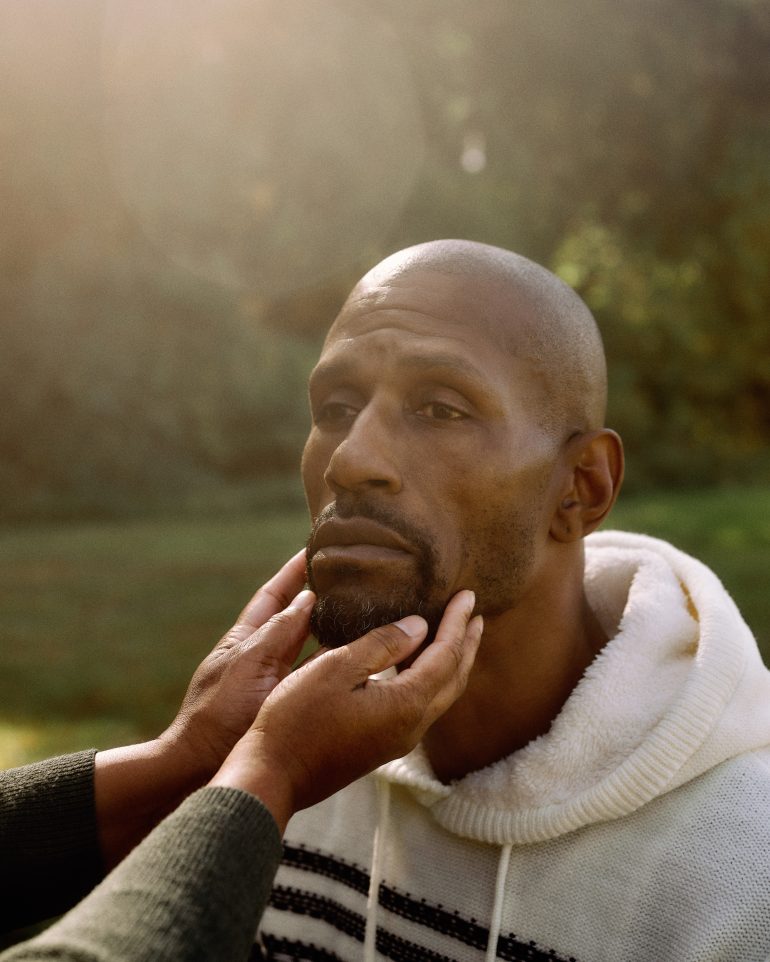 A person gently holds a mans chin outdoors as he looks ahead, wearing a white hoodie in soft sunlight.