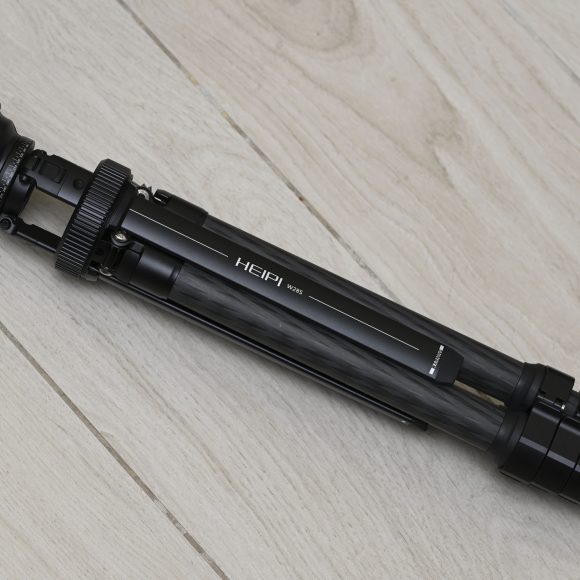 A folded black tripod labeled HEIPI 105 lying on a light wooden floor.