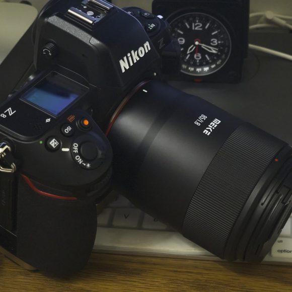 A Nikon Z camera with a large lens rests on a white keyboard near a computer and a clock.