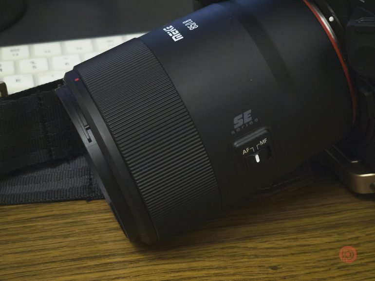 Close-up of a camera lens with focus switch, resting on a wooden surface near a keyboard.