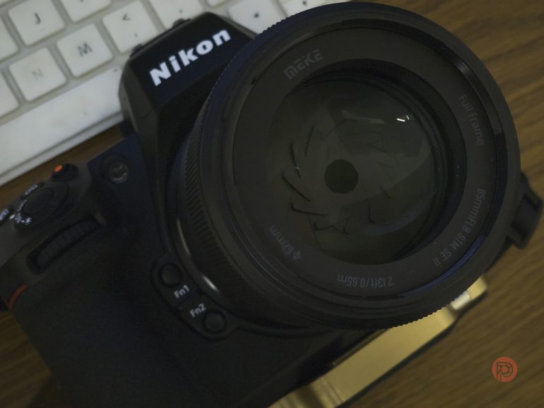 A Nikon camera with a Meike lens sits on a desk next to a white computer keyboard.