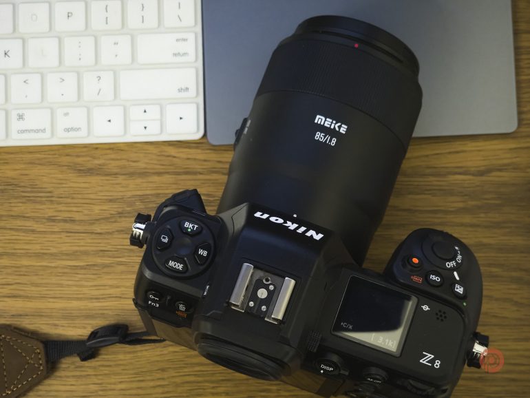 A Nikon Z8 camera with a Meike lens rests on a wooden desk beside a keyboard and a laptop.
