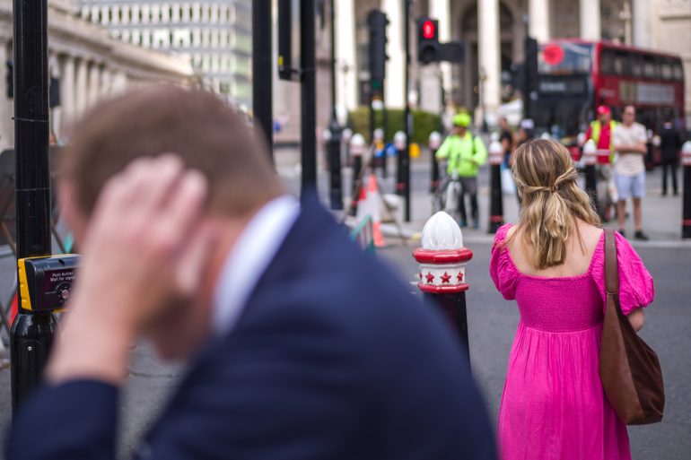 A woman in a pink dress waits at a city crosswalk; a blurred man in a suit is in the foreground.